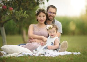 A family portrait under an apple tree featuring a mother, father, and baby girl sitting together on a blanket.