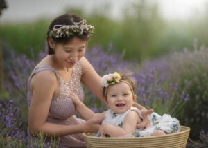 A mother in a lavender field playing with her baby girl, who sits in a basket with a floral headband and a happy smile.