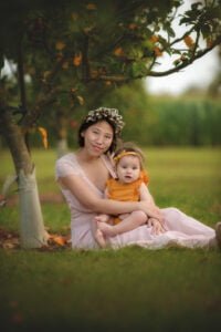Mother sitting with her baby under an apple tree, both wearing soft pastel outfits that complement the natural surroundings.