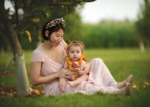 Mother and baby sitting under an apple tree, with the baby holding an apple, capturing a sweet family photography moment.