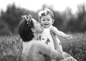 Black-and-white photo of a mother kissing her smiling baby girl in a lavender field, celebrating love and connection.
