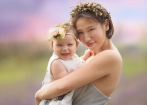 Mother smiling with her baby in her arms, both wearing floral crowns, surrounded by a colorful sunset in a lavender field.