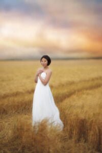 A serene woman stands in a golden wheat field, her white dress and contemplative gaze reflecting the calm majesty of the landscape around her.