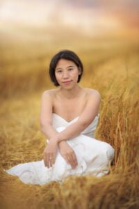 A mother in a white dress sitting in a golden wheat field, looking serene and reflective in the natural light.