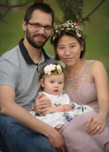 Family portrait of a father, mother, and baby sitting closely together, with floral crowns and pastel tones in a natural setting.