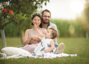 Family portrait in a meadow under an apple tree, with a baby girl reaching out toward the fruit.