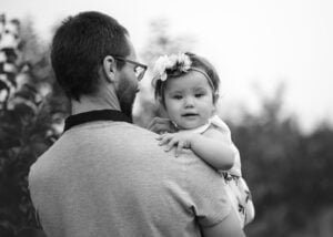 Black-and-white photo of a father holding his baby girl over his shoulder, with her floral headband adding a soft contrast.