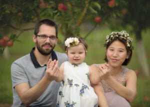 A smiling baby girl in a white floral dress, sitting with her parents who are holding her hands, under an apple tree.