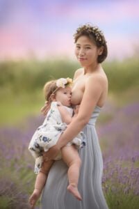 Mother breastfeeding her baby in a lavender field, wearing a floral crown and flowing dress, surrounded by serene natural beauty.