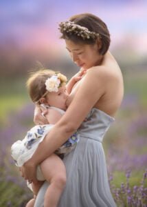Mother breastfeeding her baby in a lavender field at sunset, surrounded by soft pastel hues and floral accessories.