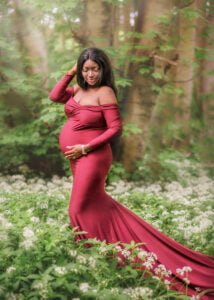 Mother-to-be in a stunning red gown contemplates the journey ahead, enveloped by a forest in bloom during her outdoor maternity photoshoot.