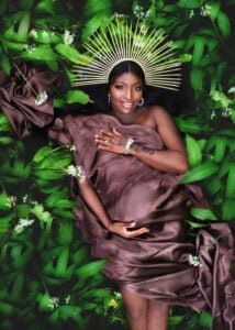 Pregnant woman wearing a celestial headpiece lying in lush green foliage during an outdoor maternity photoshoot.
