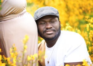 Soon-to-be father resting his head against the baby bump, surrounded by vibrant yellow flowers during an outdoor maternity photoshoot.