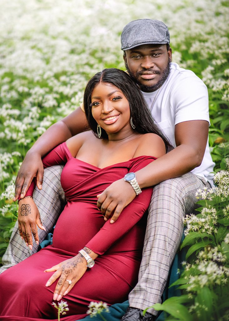 A couple cozied up amidst a field of white flowers shares a moment of contentment in their outdoor maternity photoshoot.