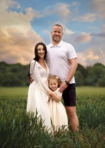 Family of three smiling during a baby bump photo shoot in a Nottingham field at sunset.