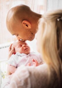 Father kissing baby’s forehead during a lifestyle newborn photoshoot at home in Nottingham, with mother holding the baby lovingly.