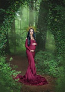 A mother-to-be stands in a mystical forest, her burgundy maternity dress and striking sunburst headpiece creating an image of serene power and grace.