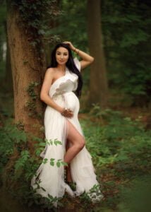 Elegant pregnant woman in a flowing white gown posing by a tree during a baby bump photo shoot in the forest.