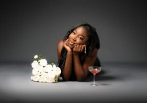 Woman lying beside white roses and a cocktail, celebrating a birthday photoshoot with joy and elegance.