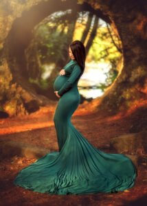 A pregnant woman in a flowing teal gown stands in a forest, with a natural arch in the background framing the scene.