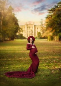 Pregnant woman in a burgundy gown posing in front of a manor during a maternity photoshoot.