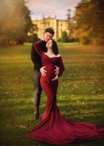 An expectant couple shares a tender moment in an autumnal park, with a majestic castle in the background.