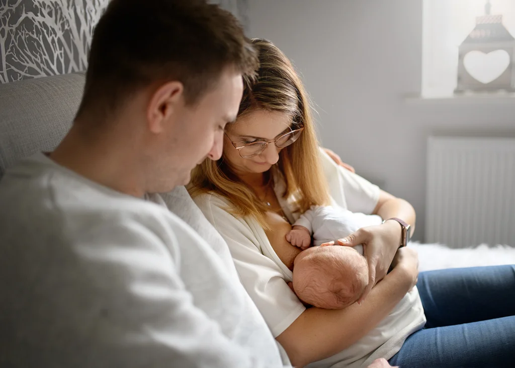 Mom breastfeeding her baby on a bed while dad lovingly watches during a newborn photoshoot at home.