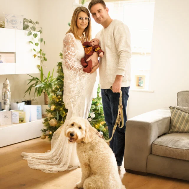 Family with their newborn baby and dog standing by a decorated Christmas tree during a newborn photoshoot at home.