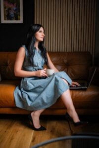 Businesswoman in a casual dress holding a coffee cup and laptop during a professional business photoshoot in a cozy workspace.