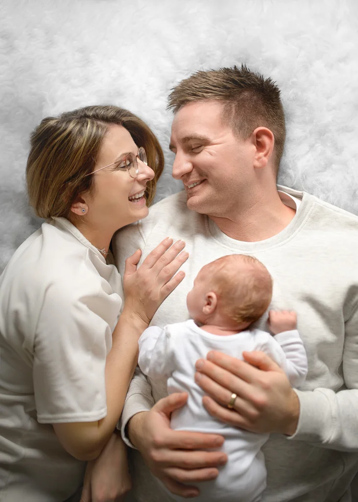 Parents smiling at each other as their newborn baby rests on dad’s chest during a newborn photoshoot at home.