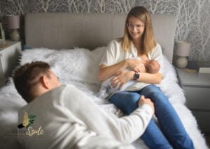 Mom breastfeeding her baby on a cozy bed during a newborn photoshoot at home while dad watches lovingly.