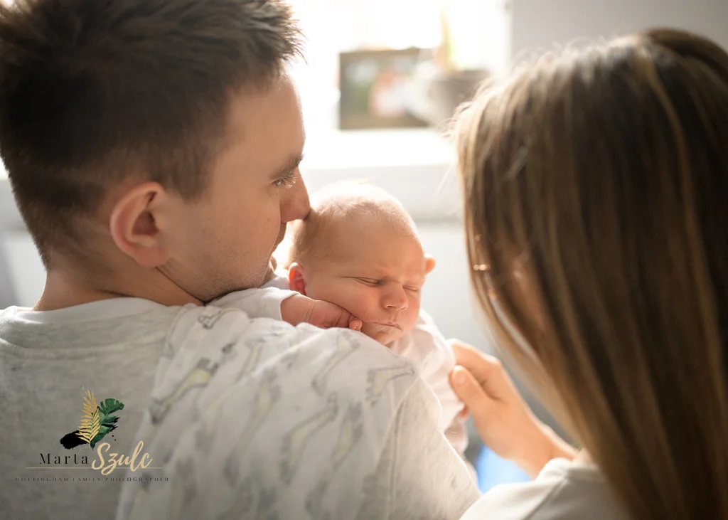 Dad holding a sleeping newborn baby as mom looks on lovingly during a newborn photoshoot at home.
