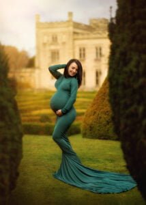 Pregnant woman in a teal maternity gown posing confidently in a manicured garden with a historic manor in the background.
