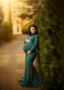 Pregnant woman in a teal dress standing by a manicured hedge during a maternity photoshoot at a historic estate.