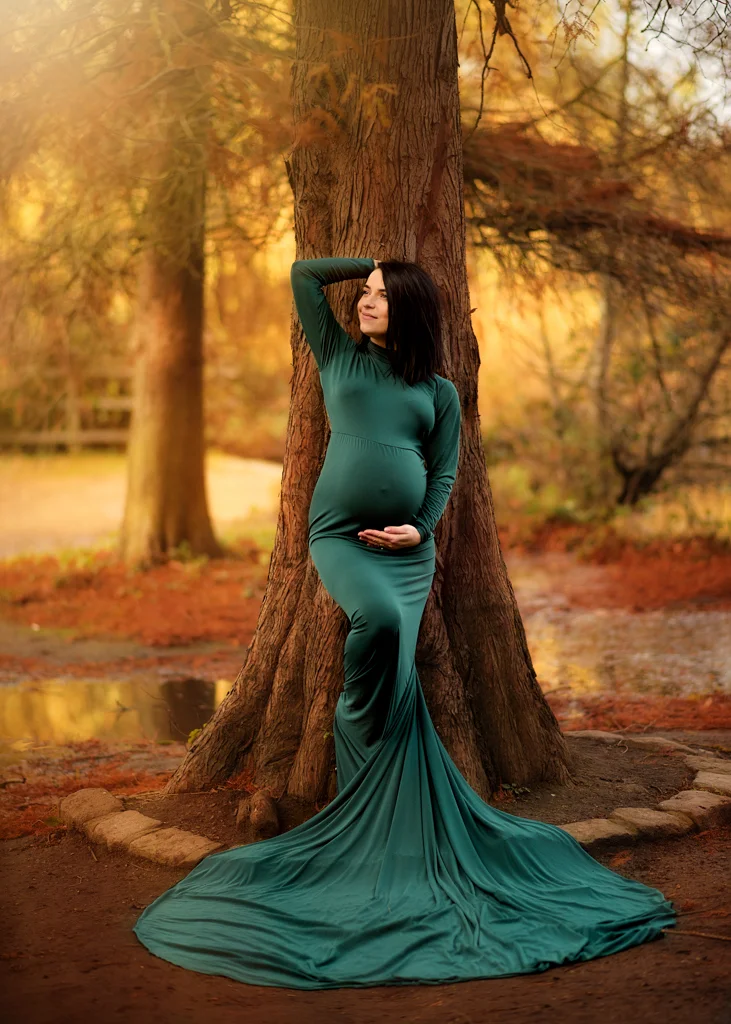 Pregnant woman in a teal maternity gown posing by a tree during a sunset maternity photoshoot.