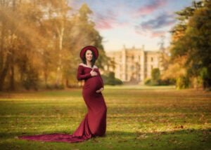 Pregnant woman in a burgundy gown smiling confidently in a sunlit field with a historic manor in the background during her maternity photoshoot.