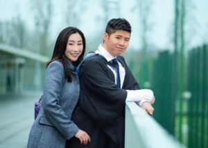 Smiling couple on graduation day at Nottingham, leaning on a railing with a backdrop of greenery and soft rain
