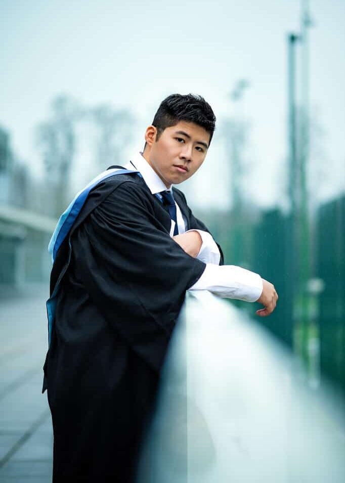 Serious young man in graduation attire at the University of Nottingham, contemplating his future on a rainy day.
