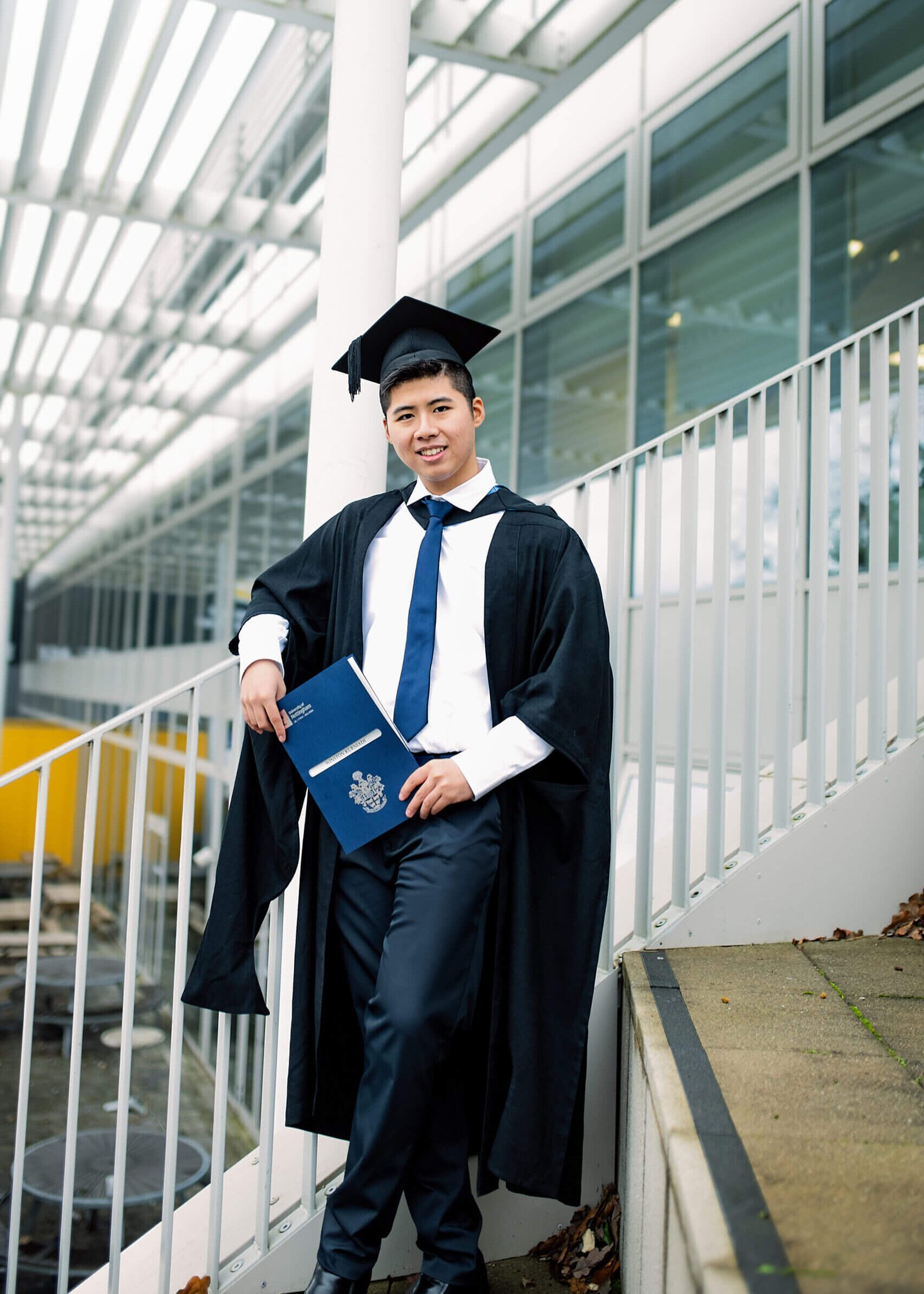 Male graduate in cap and gown holding diploma on Nottingham University campus