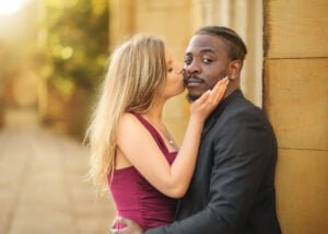 Romantic engagement photoshoot with a woman kissing her partner’s cheek against a golden architectural backdrop.
