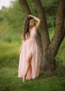 Outdoor female portrait photography of a woman in a light pink dress leaning against a tree in a natural setting.
