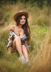 Outdoor female portrait photography of a stylish woman in a plaid shirt and tan hat, sitting in tall grass.