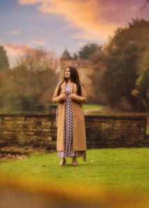 Woman in a long dress and coat posing confidently on grass with a castle in the background during her outdoor photoshoot for women.