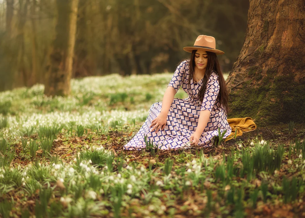Woman in a patterned dress and hat sitting under a tree, gently touching spring flowers during her outdoor photoshoot for women.