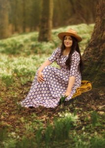 Smiling woman in a patterned dress sitting under a tree surrounded by spring blooms during her outdoor photoshoot for women.