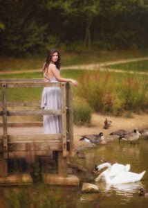 Outdoor female portrait photography of a woman in a lavender dress standing on a wooden bridge by a tranquil lake with swans.