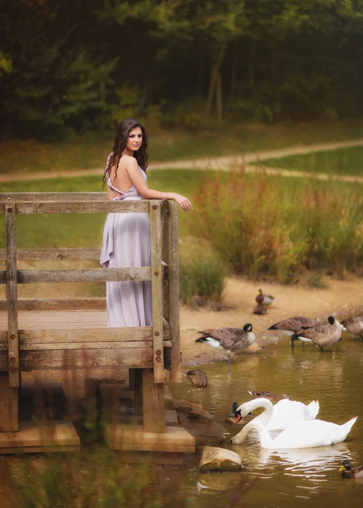 Outdoor female portrait photography of a woman in a lavender dress standing on a wooden bridge by a tranquil lake with swans.