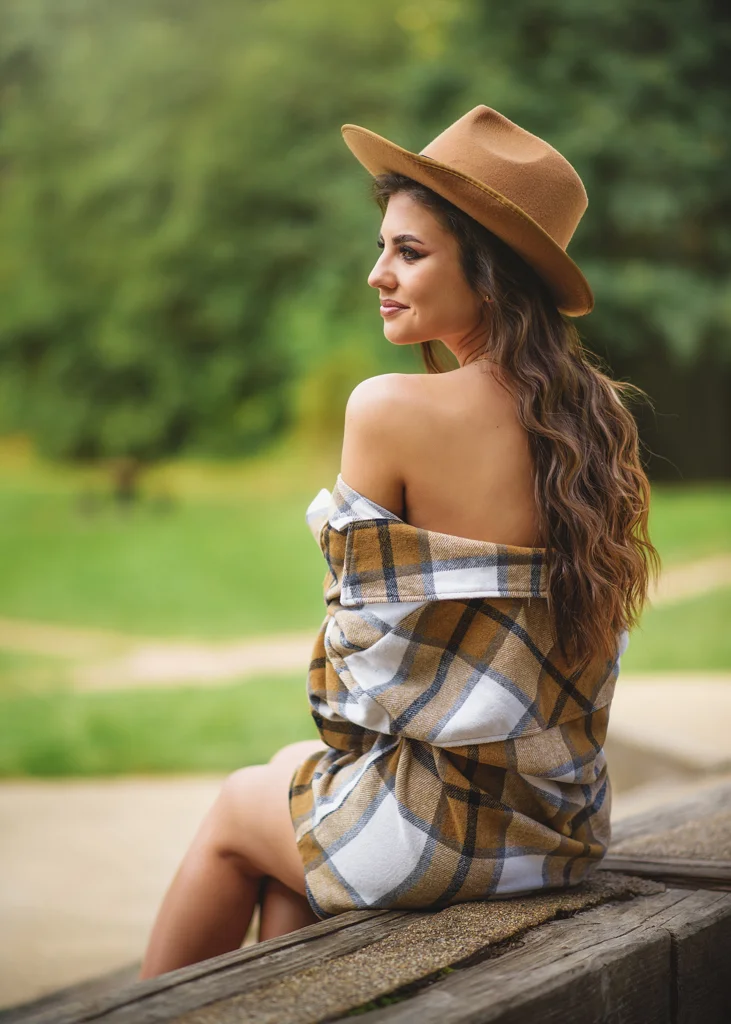 Outdoor female portrait photography of a woman in a plaid shirt and tan hat, seated on a wooden structure overlooking a green park.