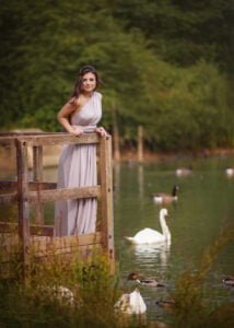 Outdoor female portrait photography of a woman in a lavender dress on a wooden bridge, overlooking swans on a calm lake.