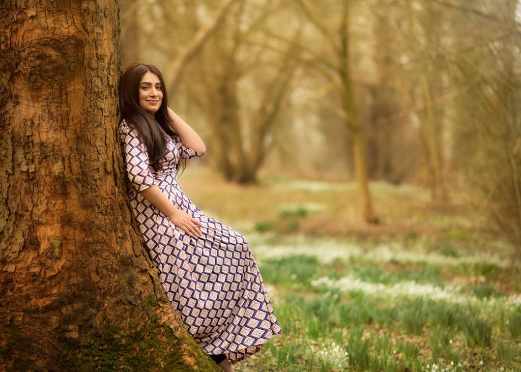 Woman in a patterned dress leaning against a tree in a springtime woodland for her outdoor birthday photoshoot.